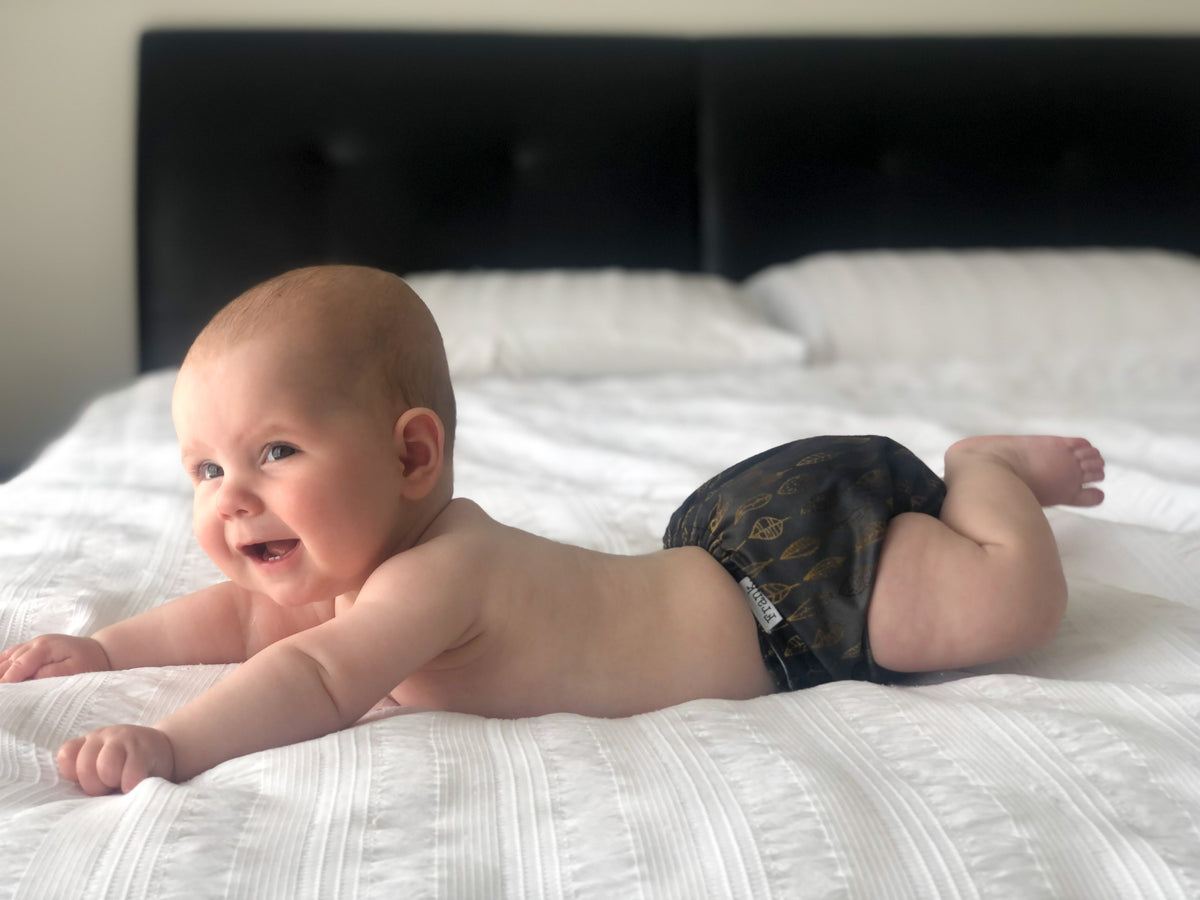 A gorgeous photo of a cute baby wearing a leaves print modern cloth nappy. The baby is stretched out on a bed and smiling. 